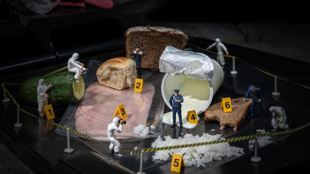 Miniaturfiguren in Schutzanzügen untersuchen Lebensmittelreste auf schwarzer Fläche, markiert mit gelben Nummernschildern, umgeben von Absperrband.