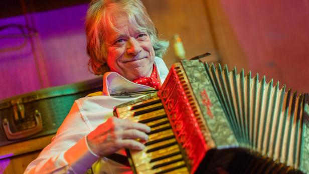 Ein älterer Mann mit weißem Hemd und rotem Halstuch spielt lächelnd ein Akkordeon.
