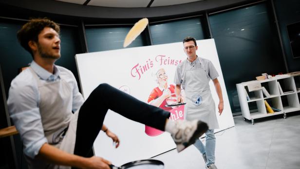 Zwei Männer werfen eine Palatschinke mit Pfannen.