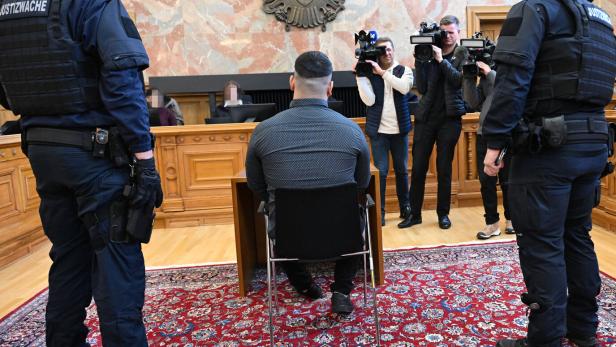 Mann sitzt mit dem Rücken zur Kamera in einem Gerichtssaal, flankiert von Justizwache