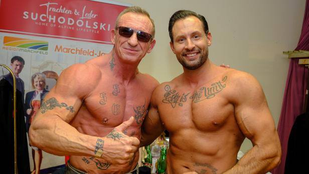 Zwei muskulöse, tätowierte Männer posieren oberkörperfrei und lächeln, einer zeigt den Daumen nach oben.