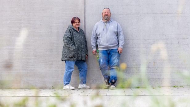 Zwei Personen stehen entspannt nebeneinander an einer grauen Betonwand, im Vordergrund unscharfe Gräser.