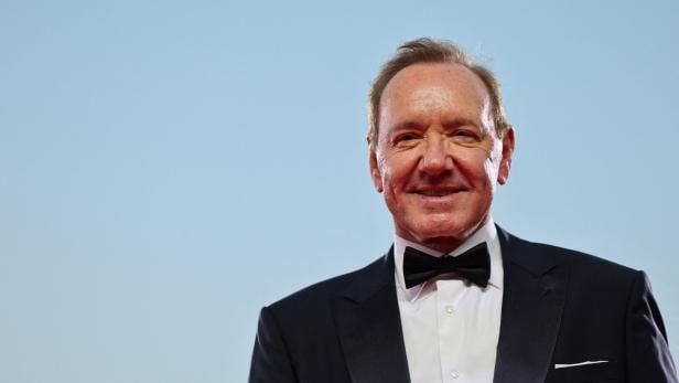 Kevin Spacey lächelt in einem dunklen Anzug mit Fliege vor hellblauem Hintergrund.