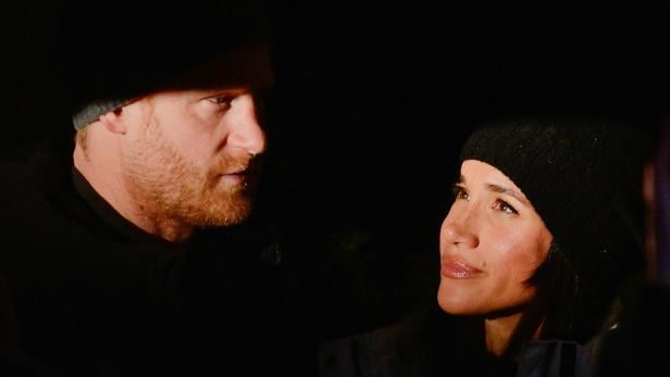 Harry und Meghan, beide mit Mützen, schauen einander im Dunkeln an.