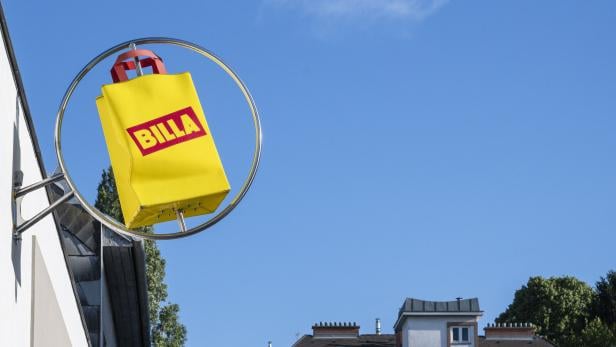 Gelbes BILLA-Schild in Form einer Einkaufstasche hängt an einer Hauswand vor blauem Himmel.
