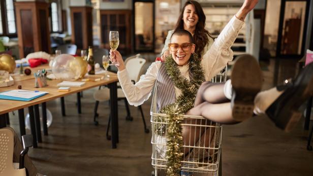 Zwei lachende Personen feiern ausgelassen, eine sitzt mit Sektglas und Girlande in einem Einkaufswagen, die andere schiebt sie.