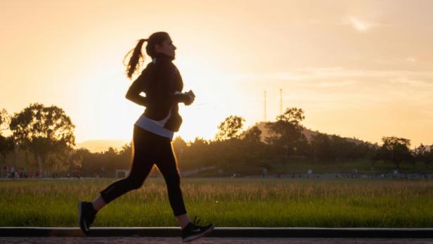 Silhouette einer joggenden Frau mit Pferdeschwanz, Sportkleidung und Laufschuhen vor Sonnenuntergang und Bäumen.