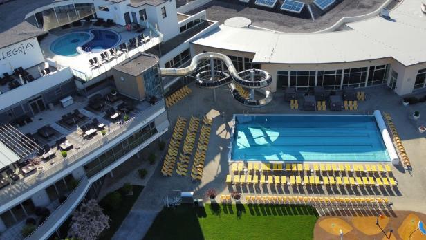 Draufsicht auf ein Schwimmbad mit Liegestühlen, Rutsche und dem Schriftzug "Allegria" auf einem Gebäude.