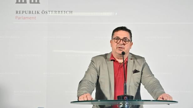 Ein Mann mit Brille spricht an einem Rednerpult vor einer Wand mit dem Logo und Schriftzug des österreichischen Parlaments.