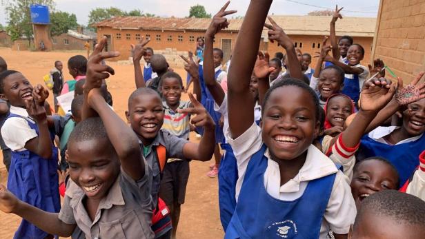 Eine Gruppe fröhlicher Schulkinder jubelt und lacht im Freien vor einem Schulgebäude.