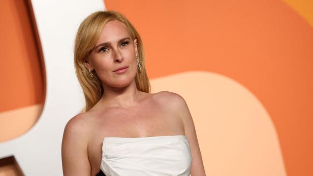 Rumer Willis mit Ohrringen trägt ein trägerloses, weiß-schwarzes Kleid vor einem orangefarbenen Hintergrund.