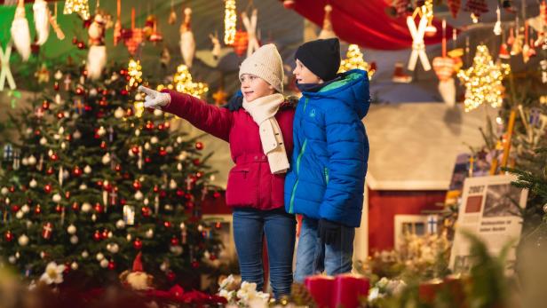 Zwei Kinder in Winterkleidung stehen vor einem Weihnachtsbaum in Hirschstetten und blicken auf einen Weihnachtsmarkt.
