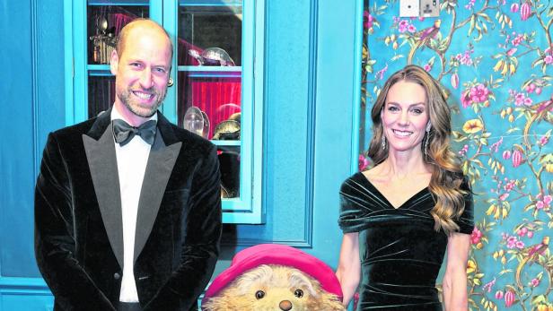 Prinz William, Kate und Paddington Bär posieren vor einer floralen Tapete.