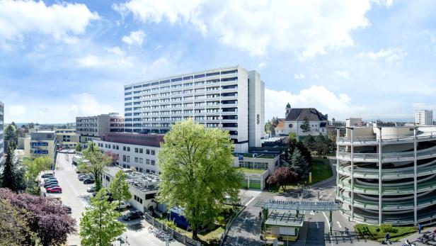 Mehrstöckiges Krankenhausgebäude mit Parkhaus, Bäumen und Zufahrt bei sonnigem Wetter.