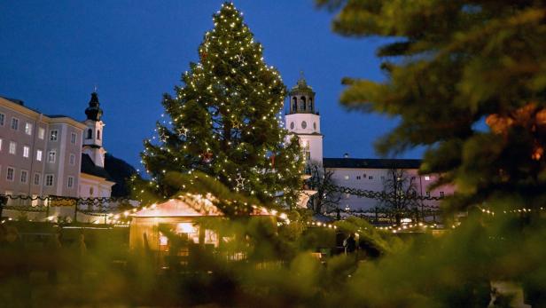 Ein Weihnachtsbaum mit Lichtern und Sternen steht auf einem Platz in Salzburg vor Gebäuden.
