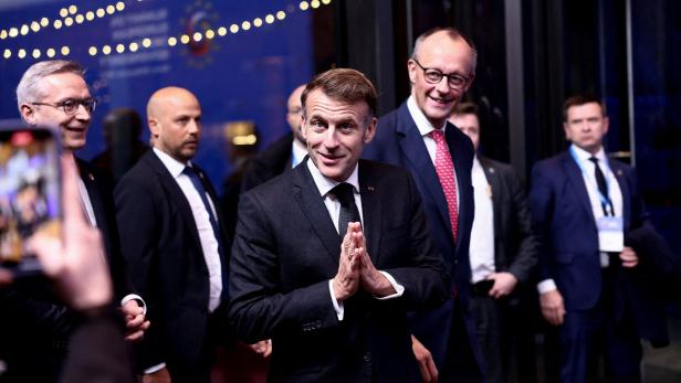 Emmanuel Macron mit gefalteten Händen, begleitet von Friedrich Merz und weiteren Personen.