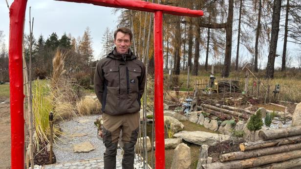 Ein Mann steht in Arbeitskleidung vor einem Garten unter einem roten Holzrahmen.