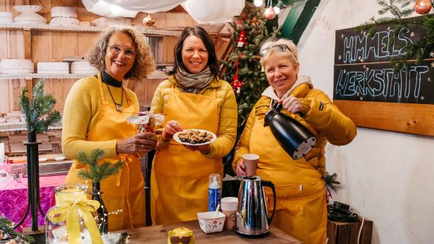 Drei Frauen in gelben Schürzen bieten Gebäck und Getränke an einem festlich geschmückten Stand an.