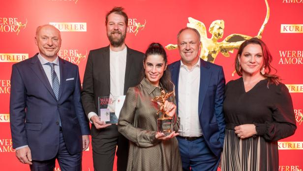 Fünf Personen in festlicher Kleidung posieren lächelnd vor rotem Hintergrund, eine Frau hält eine goldene Trophäe.