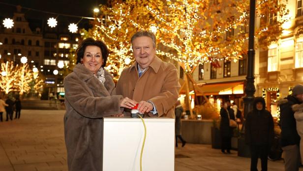 Zwei Personen drücken gemeinsam einen Knopf, im Hintergrund leuchten festlich geschmückte Bäume mit Lichterketten.