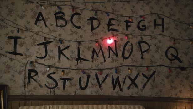 Große schwarze Buchstaben des Alphabets sind an einer Wand gemalt, darüber hängen bunte Lichterketten, ein Licht leuchtet rot.