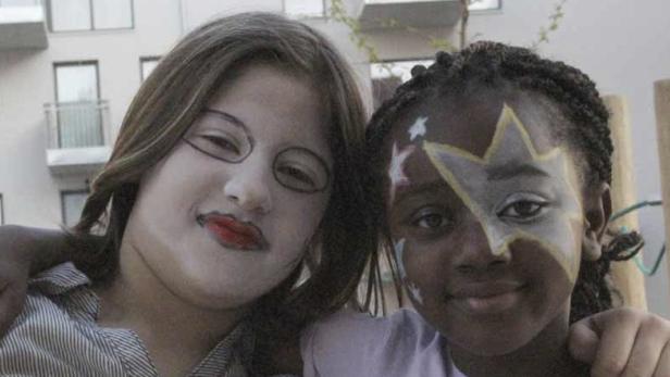 Zwei Mädchen mit Gesichtsbemalung umarmen sich vor einem Wohnhaus.