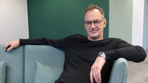 Ein Mann mit Brille und schwarzem Pullover sitzt entspannt auf einem hellgrünen Sofa vor einer grünen Wand.