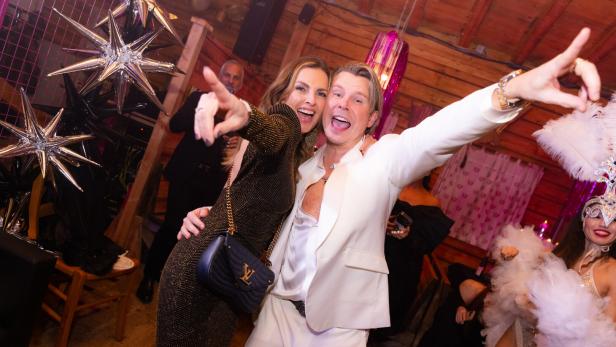 Zwei fröhliche Menschen feiern ausgelassen in eleganter Kleidung, umgeben von festlicher Dekoration und weiteren Gästen.