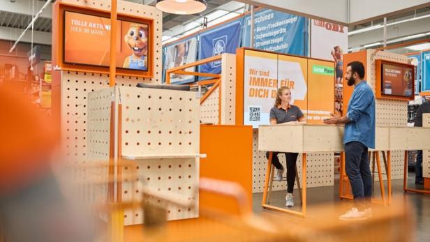 Eine Mitarbeiterin berät einen Kunden an einem Informationsstand in einem Baumarkt mit orangefarbener Einrichtung.