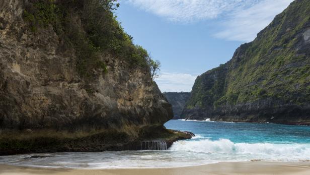 Ein Strand mit türkisfarbenem Wasser liegt zwischen bewachsenen Klippen unter einem hellblauen Himmel.