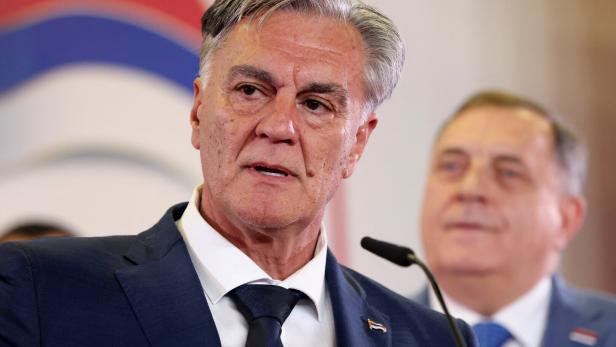 Ein älterer Mann im Anzug spricht an einem Mikrofon, im Hintergrund steht ein weiterer Mann.