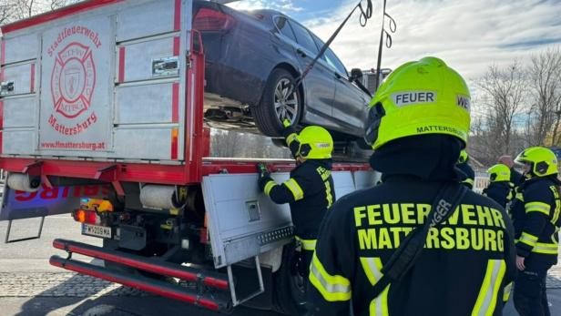 Mehrere Feuerwehrleute mit gelben Helmen verladen ein Auto mithilfe eines Krans auf einen LKW.