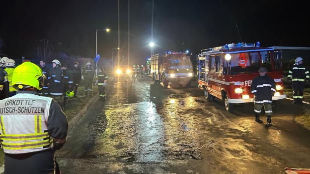 Mehrere Feuerwehrleute und Einsatzfahrzeuge stehen nachts auf einer nassen Straße, die Szene ist beleuchtet.