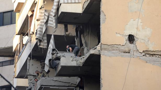 Mehrere Menschen stehen auf den zerstörten Balkonen eines schwer beschädigten Wohnhauses mit herabhängenden Vorhängen.