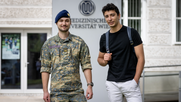 Ein Mann steht in weißer Hose, schwarzem t-Shirt und Rucksack neben einem Soldaten in Uniform, vor einem Logo der Medizinischen Universität Wien 