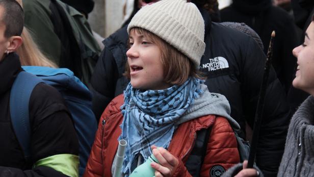 Eine junge Person mit Mütze und Schal hält eine Trinkflasche und steht inmitten einer Gruppe von Menschen.