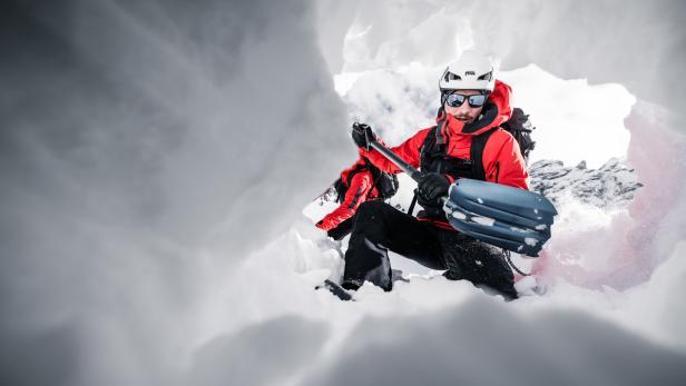 Zwei Personen in roter Winterkleidung mit Skiausrüstung in einer Schneehöhle, umgeben von Eiswänden.