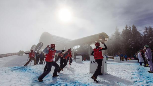 Mehrere Personen in roter Skibekleidung und Helmen stehen auf schneebedecktem Boden vor einer großen, weißen Schneekanone unter bewölktem Himmel.
