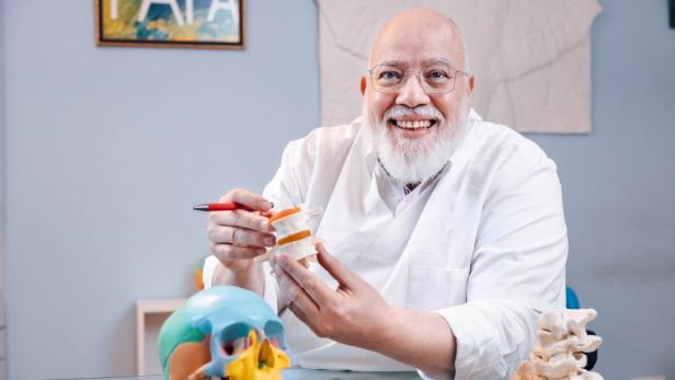 Ein Mann mit weißem Bart und Brille hält ein Knochenmodell in der Hand.