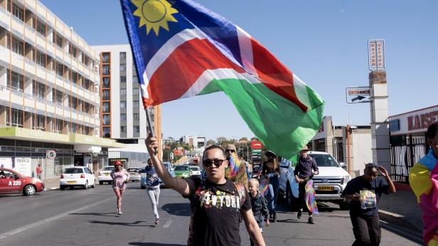 Eine Person läuft auf einer Straße und schwenkt eine große namibische Flagge, umgeben von weiteren Menschen.