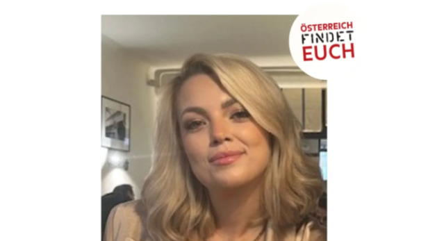 Junge Frau mit schulterlangem blondem Haar und beigem Blazer sitzt in einem Raum und lächelt leicht.