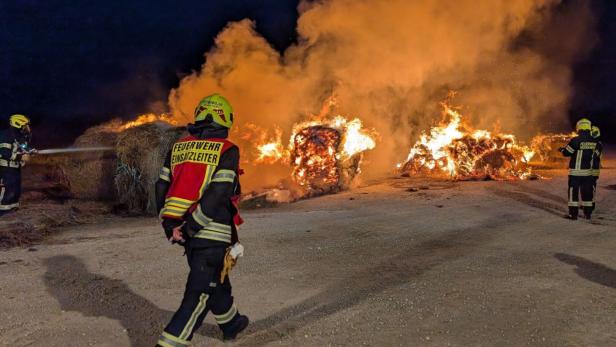 Mehrere Feuerwehrleute bekämpfen nachts einen großen Brand von brennenden Heuballen im Freien.