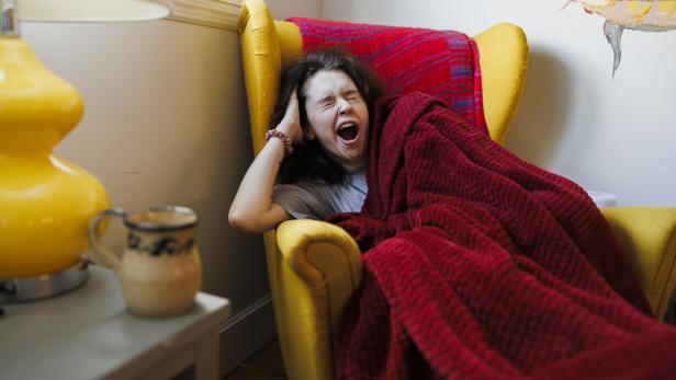 Eine Frau gähnt müde in einem gelben Sessel unter einer roten Decke.