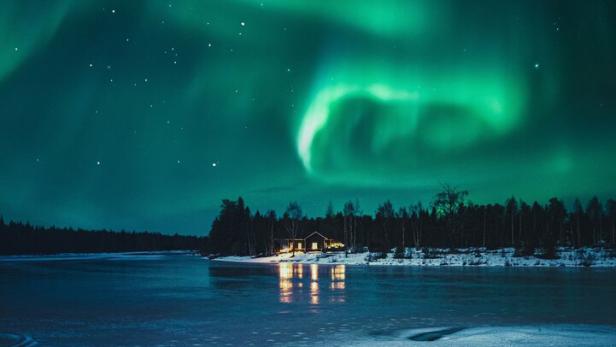 Grünes Polarlicht leuchtet am Nachthimmel über einem zugefrorenen See mit beleuchtetem Haus am Ufer.