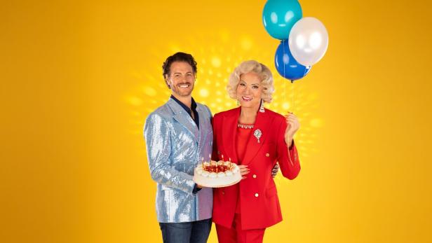 Ein Mann mit glitzerndem Sakko hält eine Torte mit Kerzen, neben einer Frau im roten Anzug mit drei Luftballons.