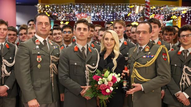 Eine Gruppe von Soldaten in Uniform steht mit einer Frau, die einen Blumenstrauß hält, vor festlicher Beleuchtung zusammen.
