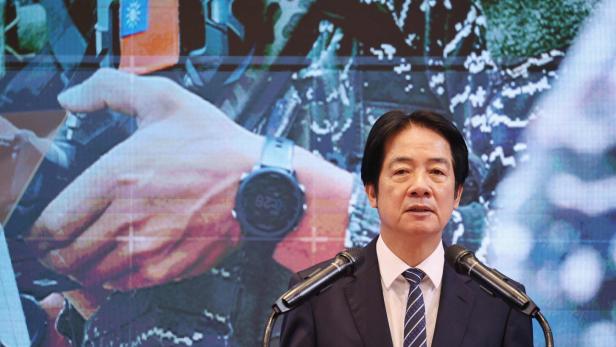 Ein Mann im Anzug spricht an zwei Mikrofonen, im Hintergrund ist eine große Nahaufnahme eines Soldaten mit Taiwan-Flagge zu sehen.