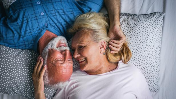 Ein älteres Paar liegt lächelnd im Bett, ihre Gesichter einander zugewandt.