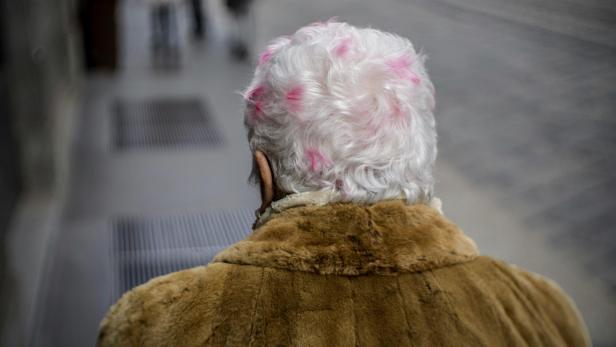 Eine ältere Person mit weißem Haar und rosa Akzenten trägt einen braunen Pelzmantel.