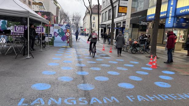 Mehrere Menschen gehen und fahren Fahrrad auf einer mit blauen Punkten und „Langsam fahren“ markierten Straße in einer Einkaufsstraße.